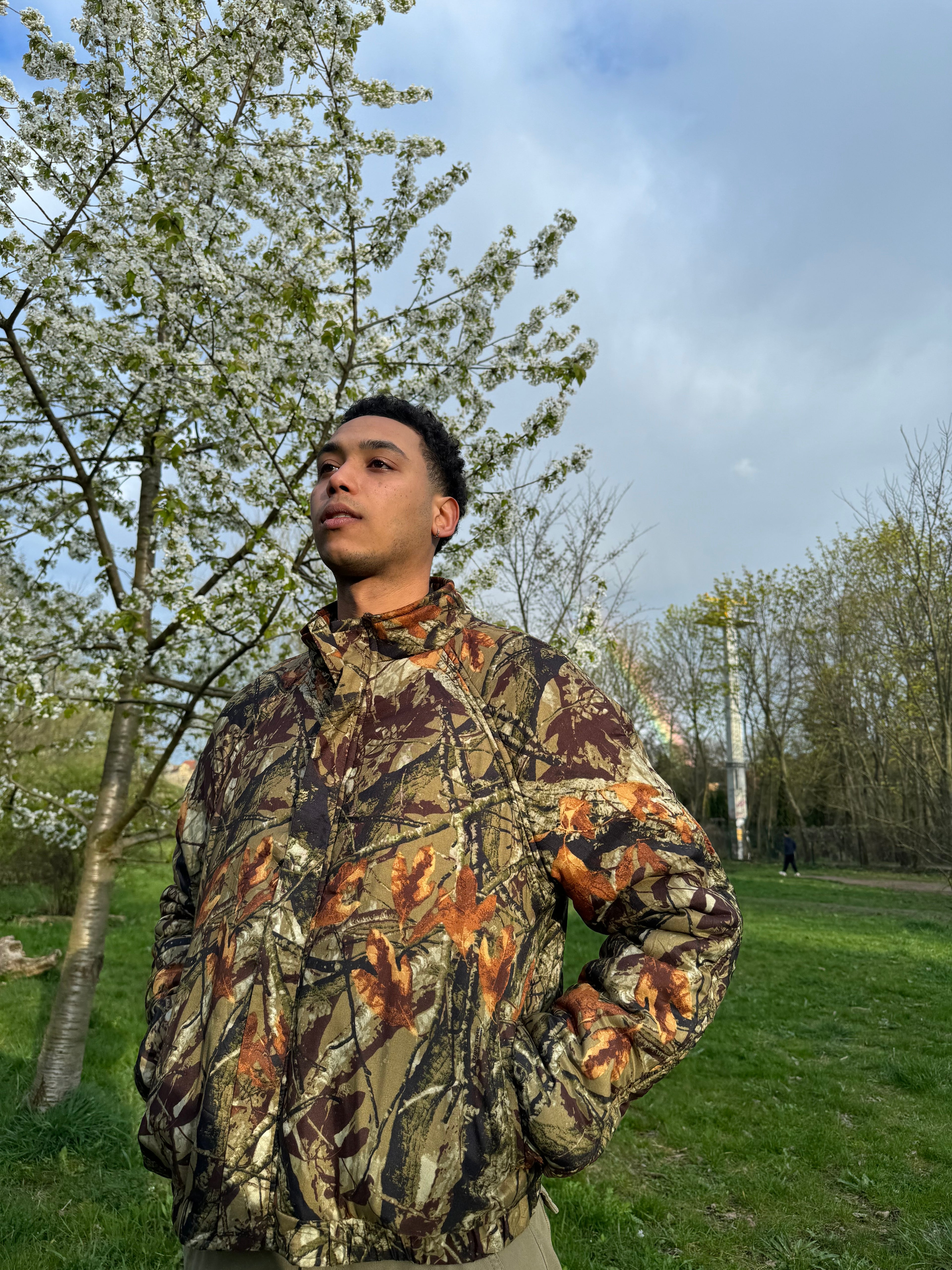 Person wearing a camouflage jacket standing outdoors with trees and grass in the background