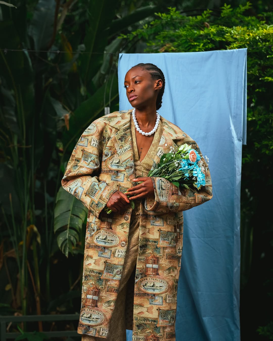 Person wearing a patterned coat holding flowers against a blue backdrop with greenery.
